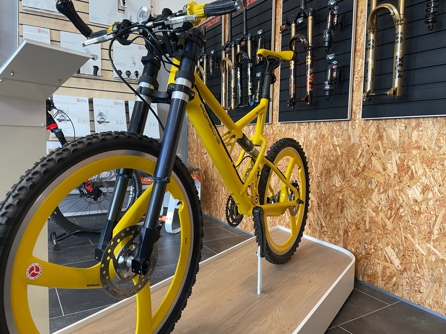 Yellow Porsche FS Evolution mountain bike from 1997 with distinctive yellow Spengle carbon three-spoke wheels, upside-down suspension fork, and Porsche branding, displayed indoors in front of a wall with various suspension forks and bike components.