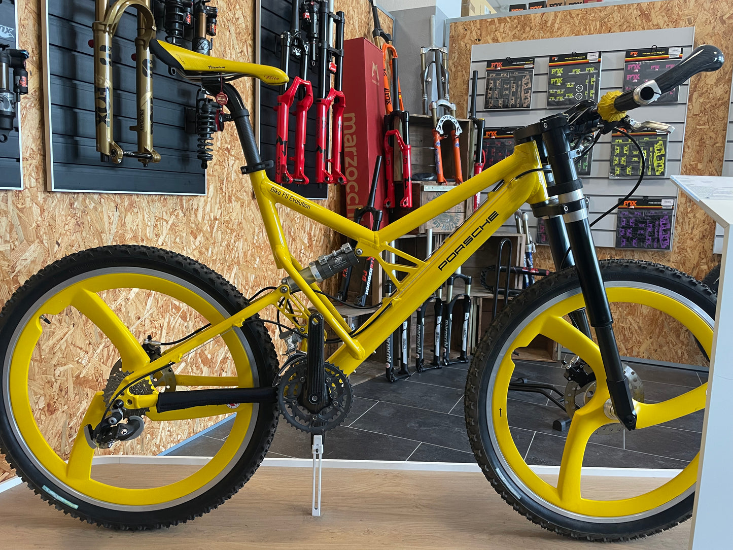 Yellow Porsche FS Evolution mountain bike from 1997 with distinctive yellow Spengle carbon three-spoke wheels, upside-down suspension fork, and Porsche branding, displayed indoors in front of a wall with various suspension forks and bike components.