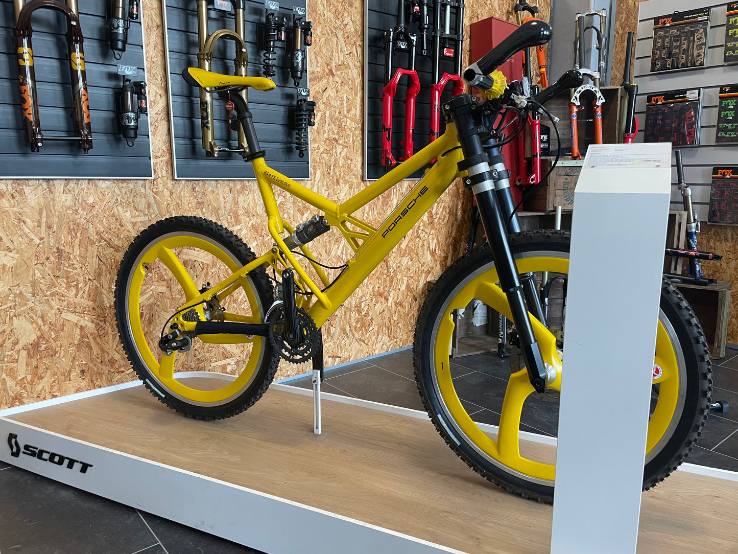 Yellow Porsche FS Evolution mountain bike from 1997 with distinctive yellow Spengle carbon three-spoke wheels, Sachs Plasma drivetrain, Hope hydraulic brakes, and an upside-down suspension fork, displayed on a Scott-branded platform in front of a wall with various bike forks and shocks.