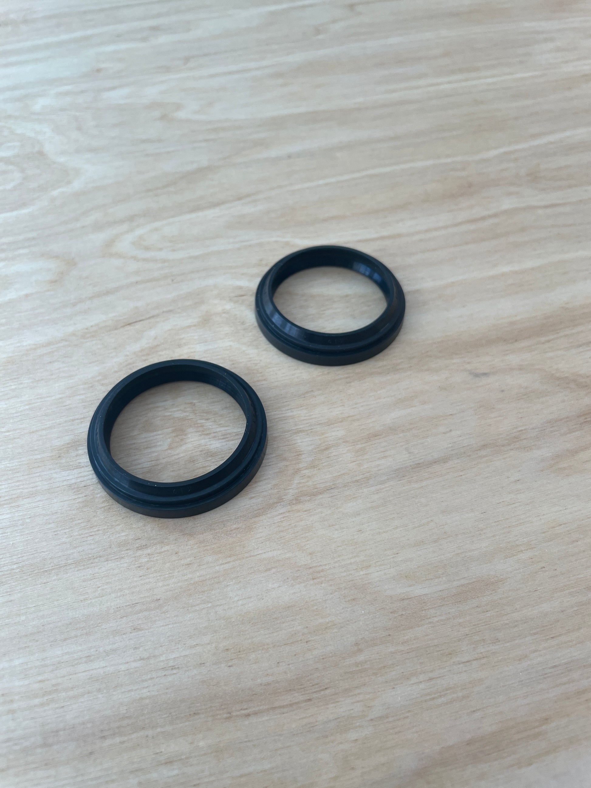 Two black Votec fork dust wipers (seals) for the lower part of the suspension fork, suitable for almost all Votec models, lying on a light wooden surface.
