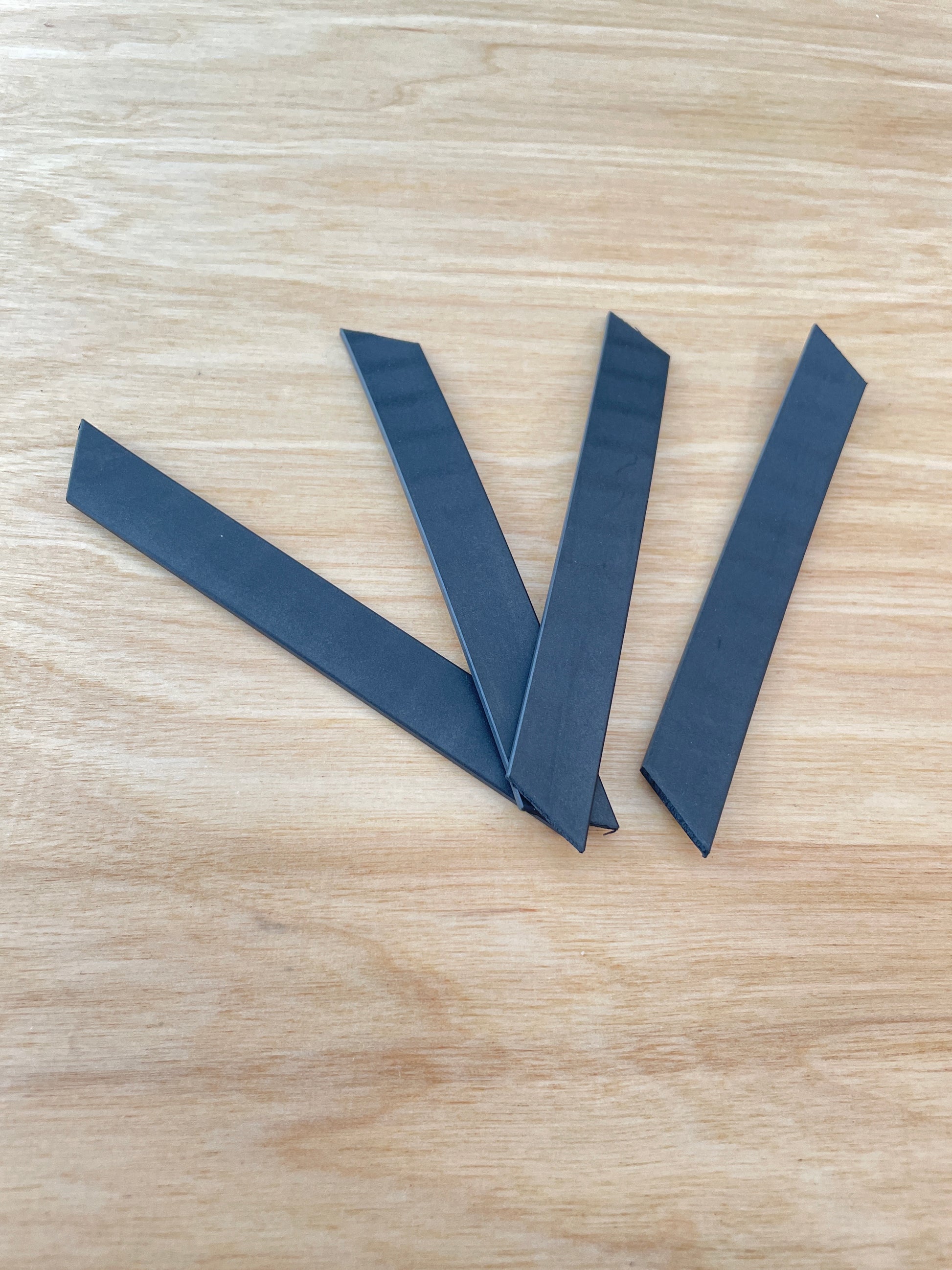 Four black, elongated carbon guide strips from Votec are arranged in a fan shape on a light wooden surface. Each strip is rectangular with one end cut at an angle, used as guide strips for the lower parts of a bicycle fork.